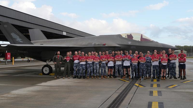 Military personnel in matching shirts stand before an aircraft.