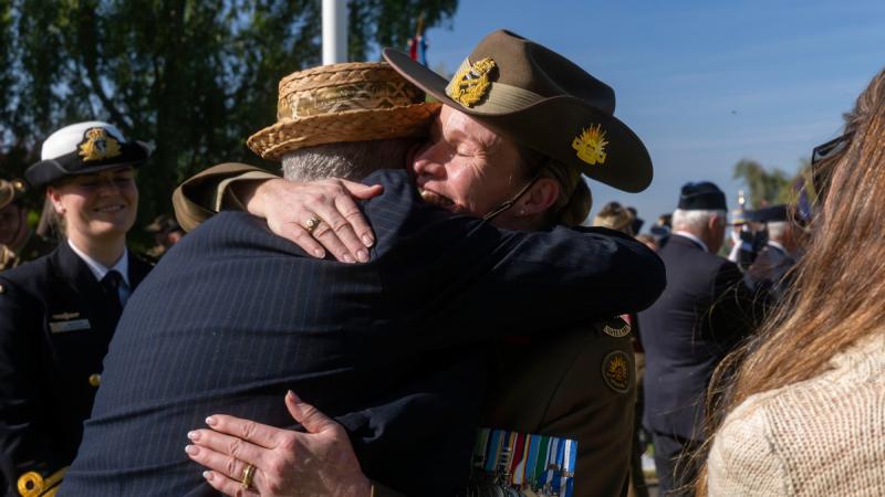 A woman in uniform hugs a man in a blazer and hat outdoors in a crowded area.