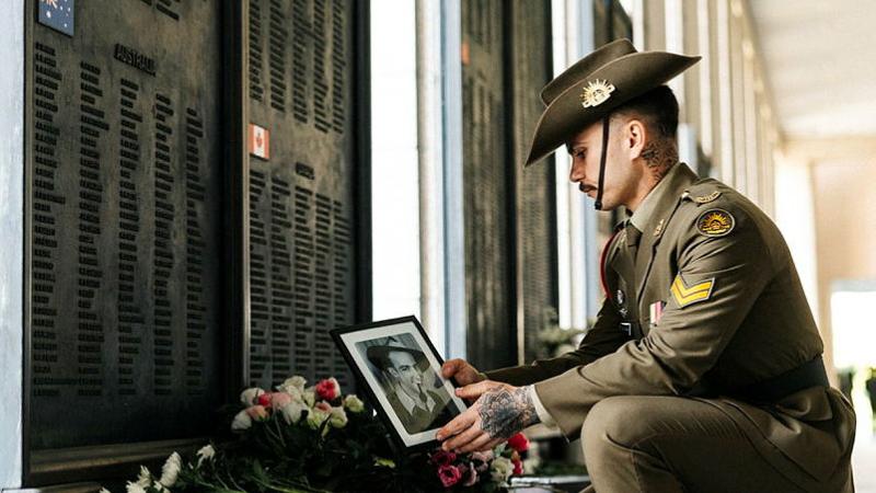 A soldier crouches at a wall  of remembrance, holding a photograph of a relative.