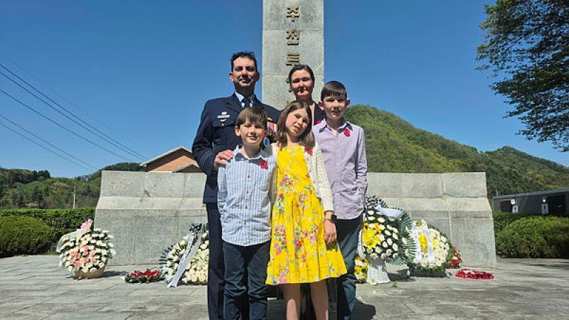 A family of two adults and three children stands in front of a stone memorial decorated with floral wreaths.