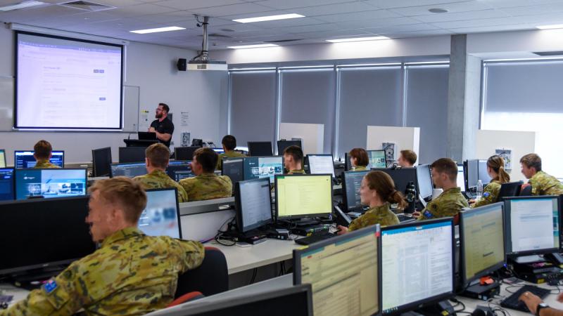 People in uniform sit at rows of desks using computers in a classroom while a man speaks at the front of the room.