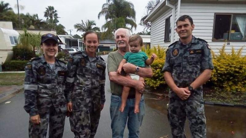 Four adults stand side by side in a wet caravan park, with one adult holding a small child.