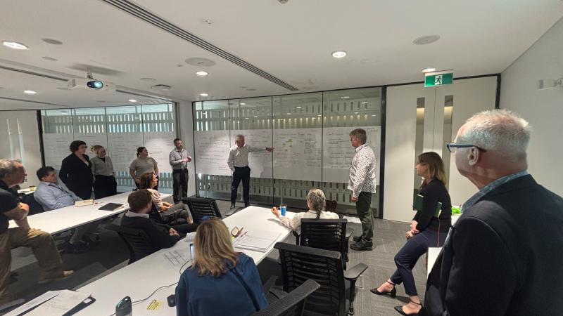 Numerous people in a classroom setting looking at writing on a whiteboard.