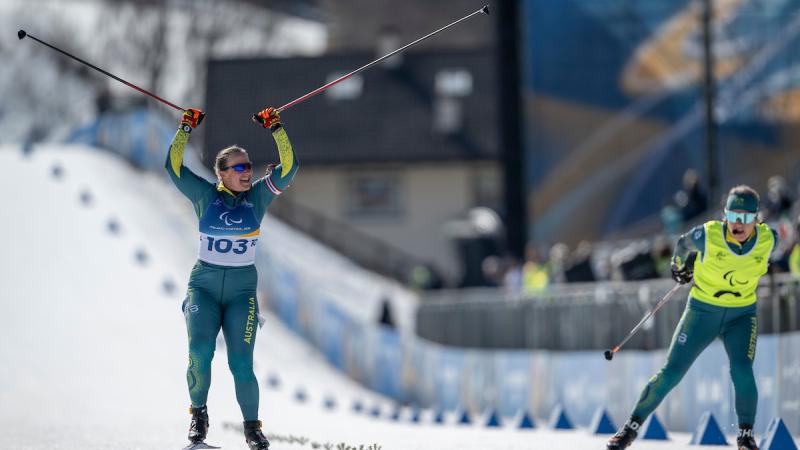 Nay Paralympian on skis and with poles in the air accompanied by her seeing guide.