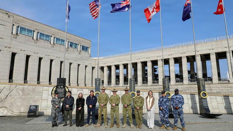 Defence personnel stand in front of flags and a building on a clear day.