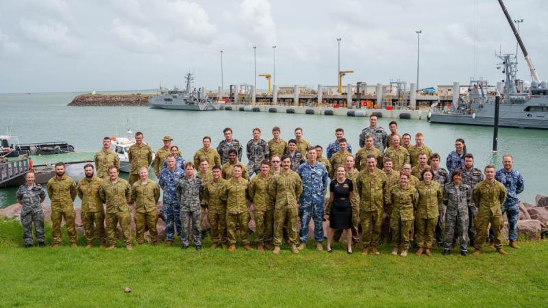 A large group of uniformed people stand on grass near water, with Navy vessels docked on the opposite shore.