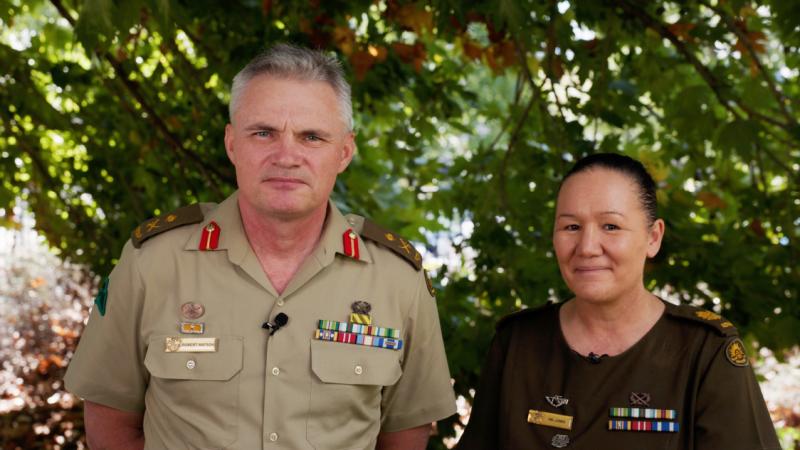 A man and a woman in Army uniforms stand next to each other under a tree.