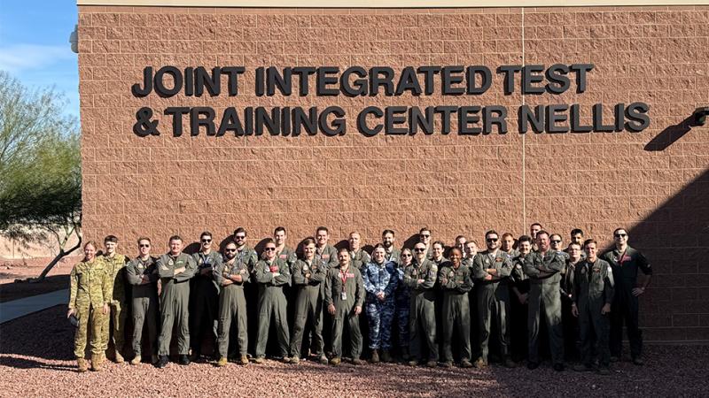 A large group of military personnel stand before the Joint Integrated Test & Training Center Nellis building.