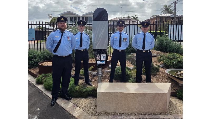 Four military men stand beside a propeller blade in an outside garden.