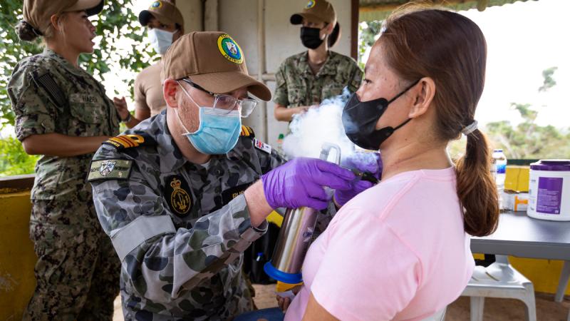 A man in Navy uniform and wearing a mask provide medical treatment to a woman.