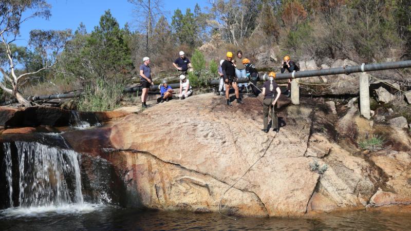 A group of veterans watch an instructor help a veteran to climb down a cliff.