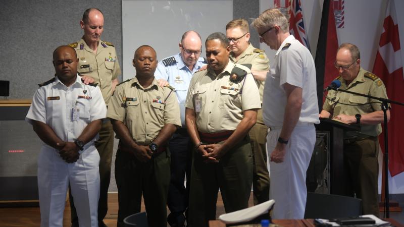 Men in uniform stand indoors with heads bowed and eyes closed, with hands resting on the shoulders of those in front.