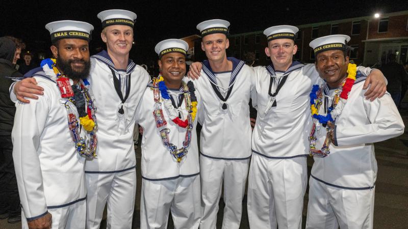 Six men in white sailor uniforms stand with their arms over each other’s shoulders, three wearing lolly leis.