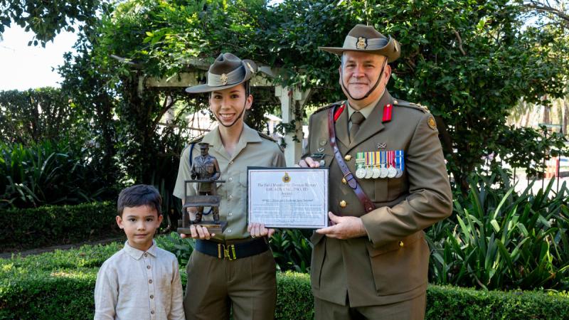 A boy stands with a woman and man in Army uniform with the women holding a trophy and certificate.