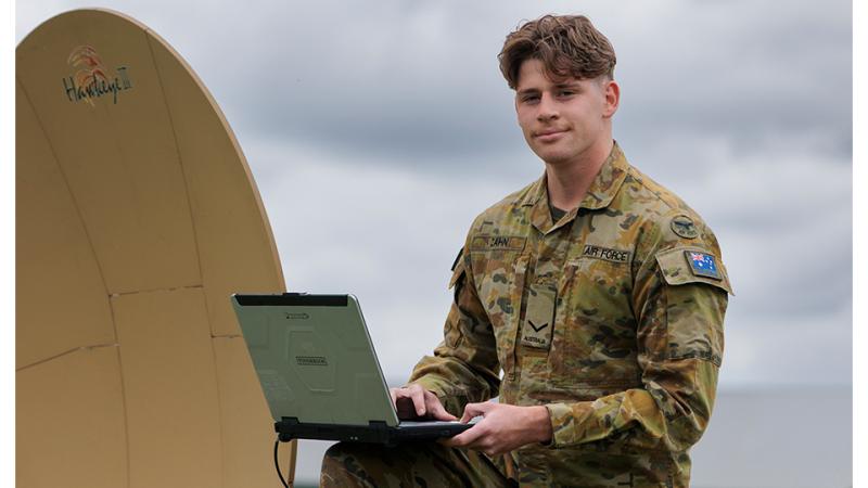 A military man kneels with a laptop open on his knee and a communications satellite dish beside him.