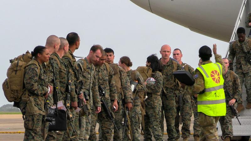 People in uniform walk down aircraft stairs onto a tarmac while others stand nearby.