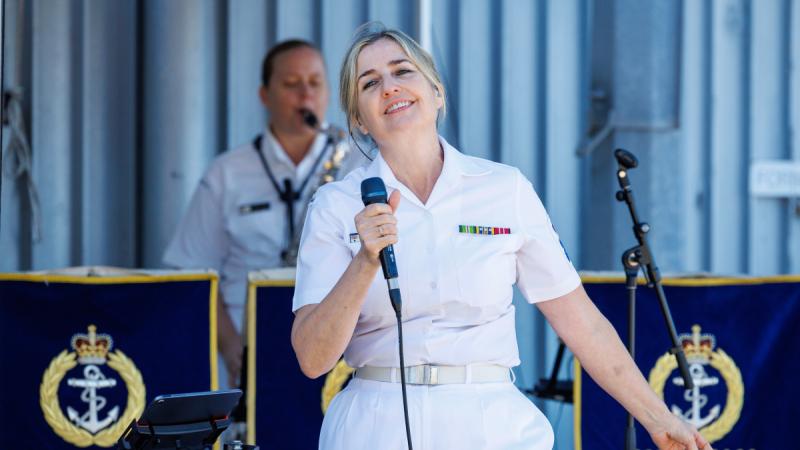 A woman in Navy uniform sings into a microphone, with music stands and a saxophonist behind her.