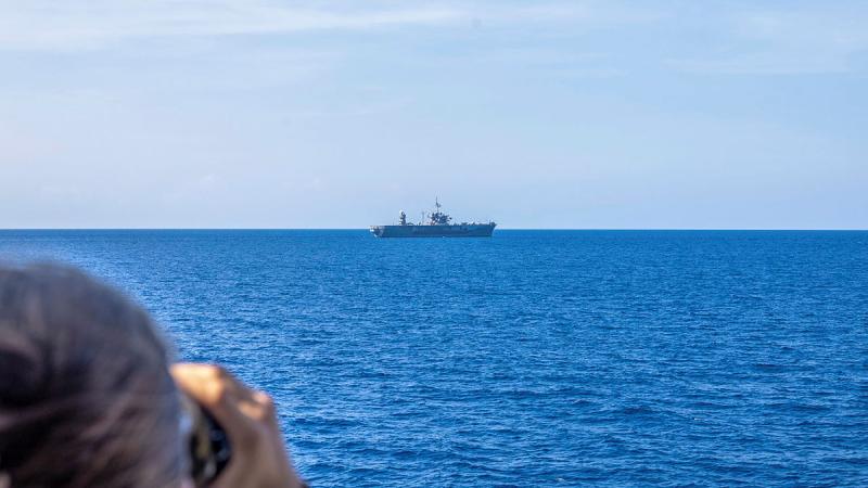 A sailor with binoculars looks out to sea to a ship in the distance.