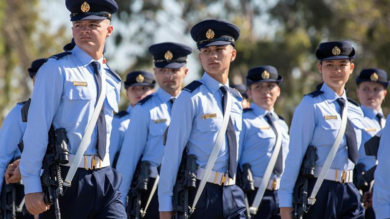 Ait Force graduates march in a group on a sunny day with trees in the background.