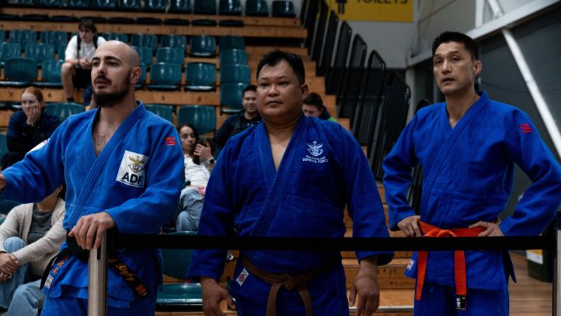 Three men in blue judo gi stand side by side watching a match, with sparsely filled bleachers behind them.