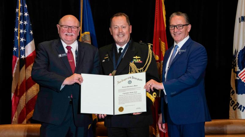Navy Commodore David Frost is holding his award and is flanked by two men.