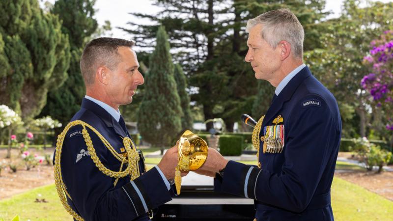Two men in formal Air Force uniforms holding a sword, a formal garden in the background.