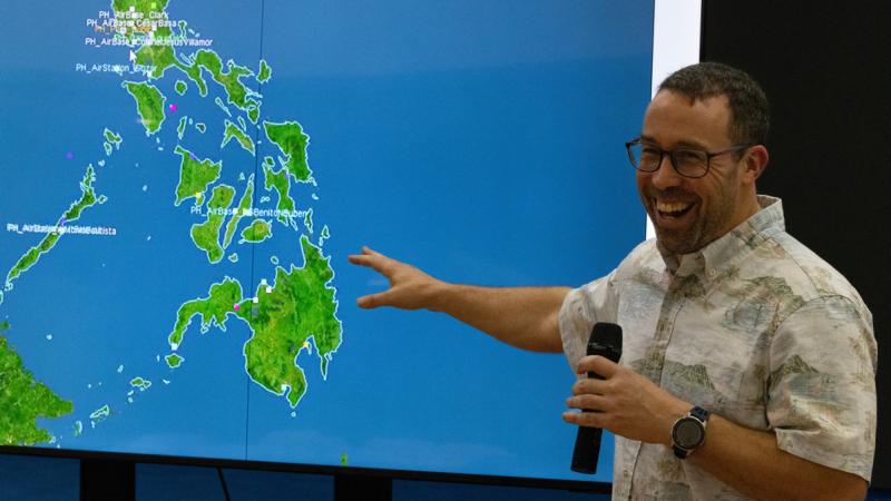 A man in a casual shirt speaks into a handheld microphone and points to a map on a screen.