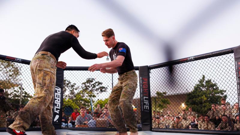 Two men wearing camouflage pants and black shirts reach towards one another in a ring.
