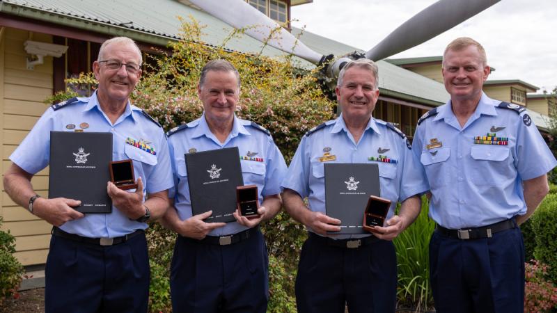 Four men in uniform stand outside a building in front of a garden with an aircraft propeller, three holding presentation boxes and black folders.