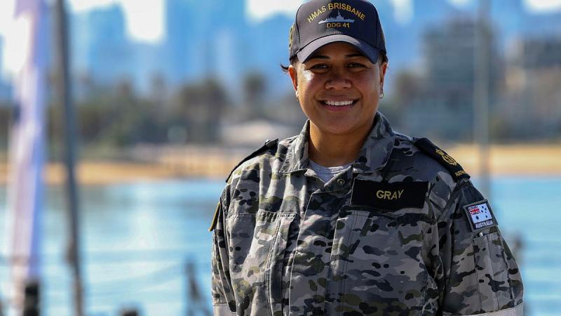 A sailor stands in front of a city skyline and beach in her Navy uniform.