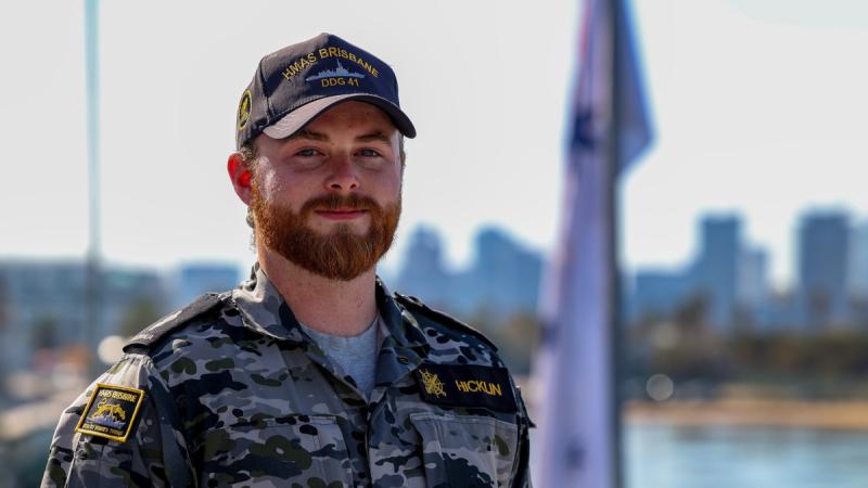 A bearded man in Navy uniform stands outdoors with water in the background.