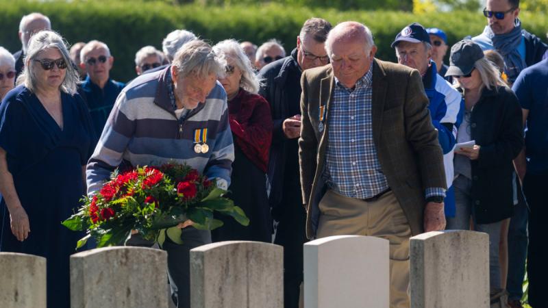 A man places a wreath on a headstone while another man pays his respects.