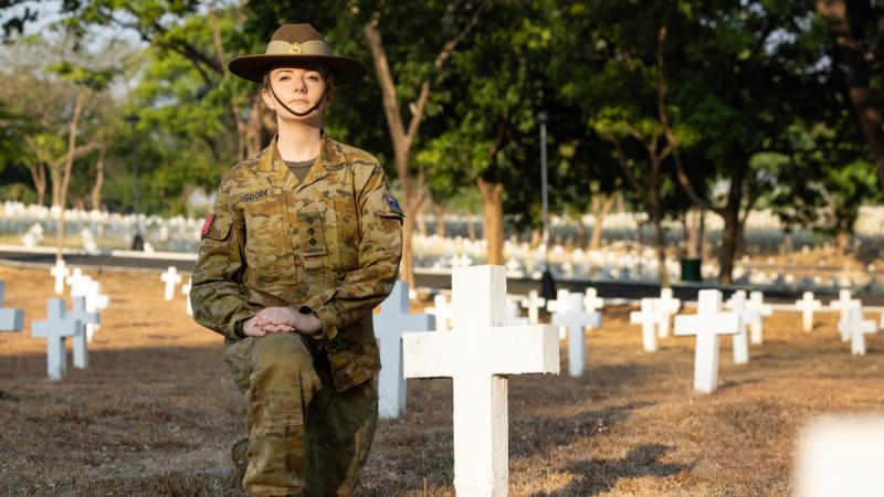 A woman in uniform kneels beside a white cross among rows of white crosses in a cemetery.