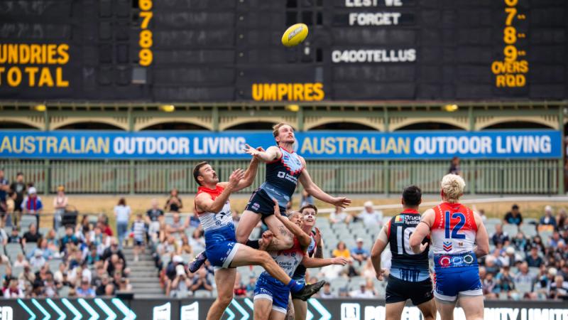 Men in AFL uniforms leap for a high ball as spectators watch from stadium seating.