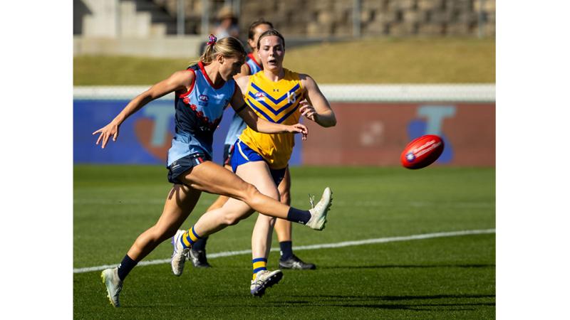 A woman in a blue AFL uniform kicks a ball as a woman in a yellow AFL uniform runs towards her.