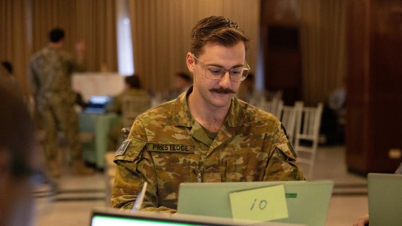 A uniformed man works on a laptop at a table, with other people partially visible at the same workspace.