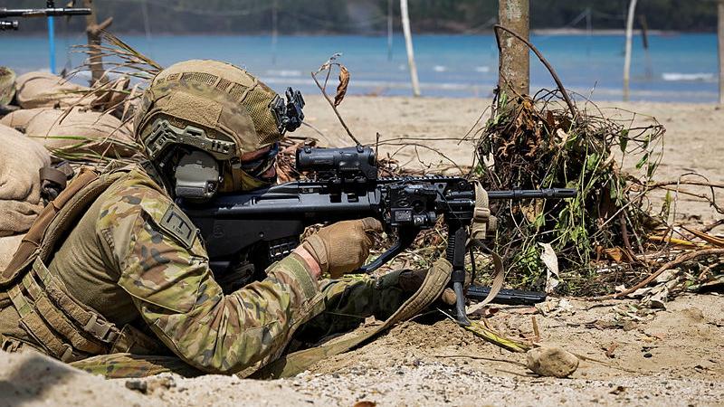 A soldier lays in the sand taking aim with a firearm towards a beach.