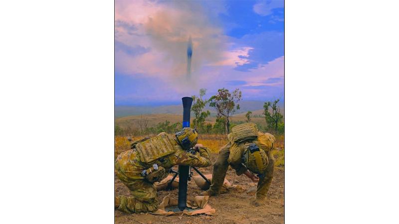 Two soldiers in camouflage uniforms and helmets crouch beside a mortar as a shell exits the tube.