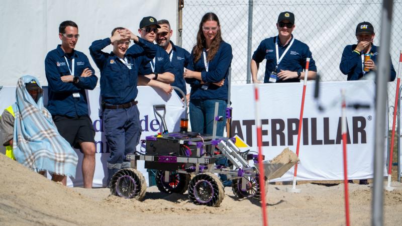The Robotics and Autonomous Systems team stand around on the sand arena operating their sub‑60kg semi‑autonomous rover. 