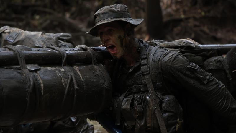 A man covered in mud carries a long mud-covered log over his shoulder in the jungle, mouth open.