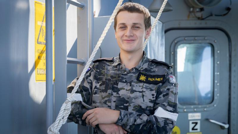 A portrait of Combat Systems Operator Seaman Eden Holmes standing at the bridge wing of HMAS Ballarat, leaning against a ladder smiling in Navy uniform.