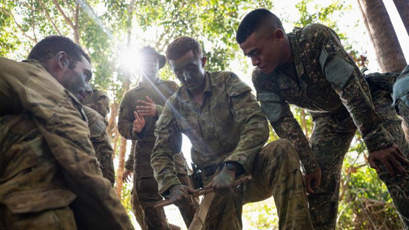 A group of Army personnel gather around a man in Army uniform rubbing two sticks together to start a fire.