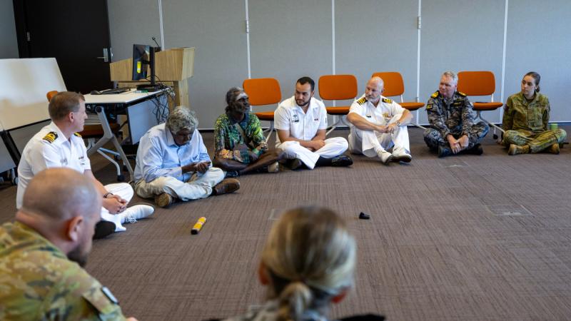 People sit cross‑legged on the floor in a circle in a room, with a message stick placed in front of one person.