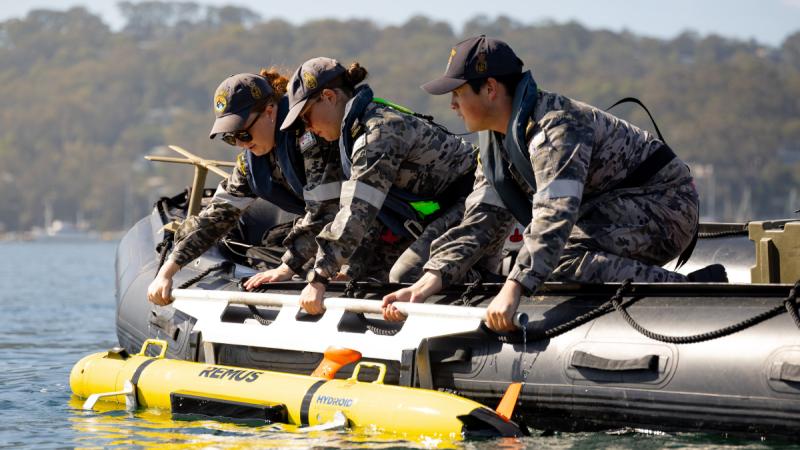Three people in Navy uniform on an inflatable boat in the water lifting out a yellow underwater vehicle.