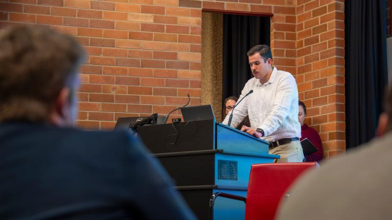 A man stands at a lectern speaking to a seated audience.