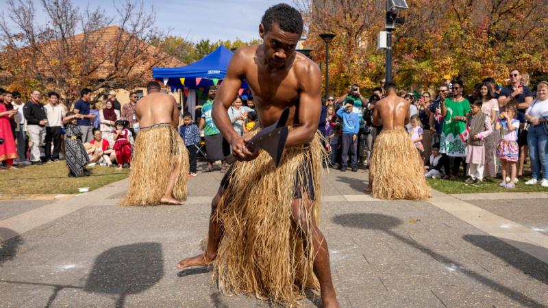 A man in a grass skirt holds a long object, while two other men in grass skirts do the same while facing a different direction as an audience watches.