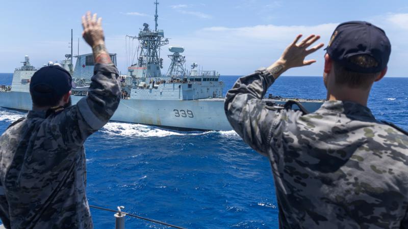 Two people in camouflage uniforms and caps wave from behind a railing toward a nearby ship.