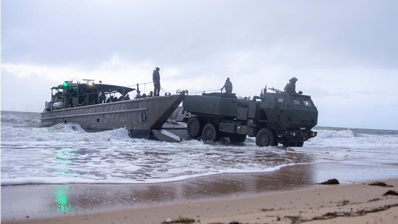 A military vehicle drives from a small vessel onto a beach.