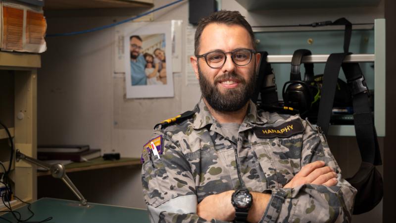 A man in uniform and glasses at a fold-down desk with his arms crossed, a photo stuck on the wall behind him.