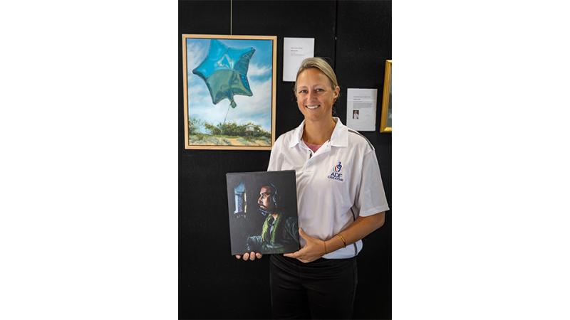 A woman in a white polo shirt stands next to one painting and holds another.